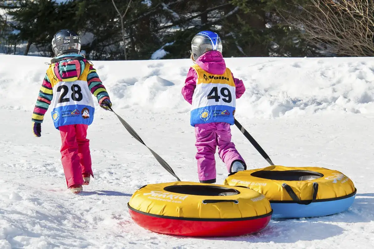 Dzieci w kolorowych kurtkach ciągną dmuchane koła po śniegu na czarnej górze snowpark.