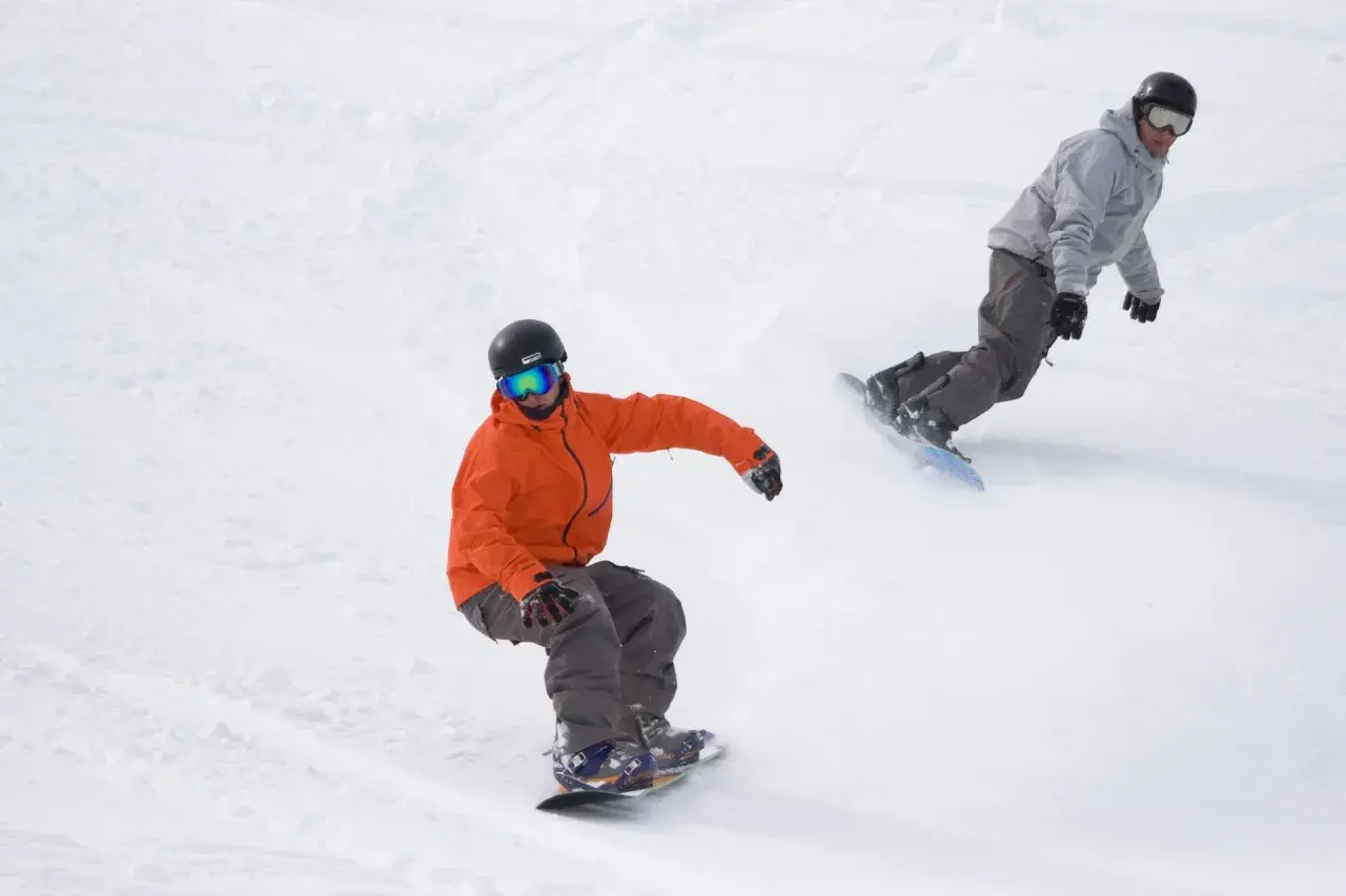 Dwóch snowboardzistów na stoku. Jeden w pomarańczowej kurtce, drugi w szarej. Uczą się, jak skręcać na snowboardzie, tworząc zaspy śniegu.